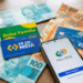 Cédulas de reais, cartões Bolsa Família e Pé-de-Meia com smartphone exibindo app Meu INSS sobre mesa de madeira.