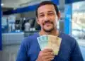 Homem brasileiro sorrindo, segurando cédulas de R$ 100 e R$ 50, representando os pagamentos de R$ 406, R$ 675 e R$ 946 que serão liberados em 15 de junho.