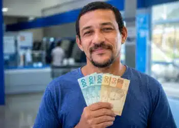 Homem brasileiro sorrindo, segurando cédulas de R$ 100 e R$ 50, representando os pagamentos de R$ 406, R$ 675 e R$ 946 que serão liberados em 15 de junho.