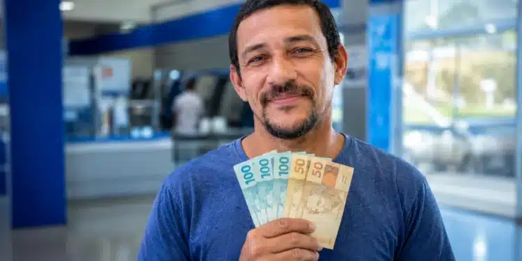 Homem brasileiro sorrindo, segurando cédulas de R$ 100 e R$ 50, representando os pagamentos de R$ 406, R$ 675 e R$ 946 que serão liberados em 15 de junho.