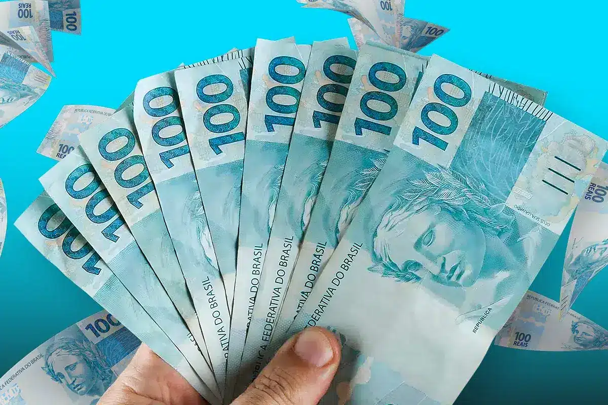 Mão segurando várias cédulas de 100 reais representando o pagamento das parcelas do incentivo de frequência do Pé-de-Meia em 2026.