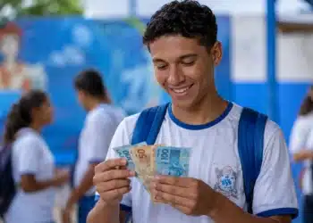 Estudante brasileiro de ensino médio segurando cédulas de reais em um pátio escolar, simbolizando recebimento de R$ 1.400 do Pé-de-Meia