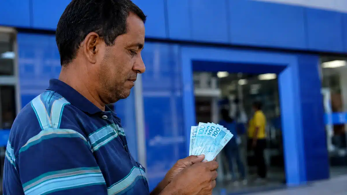 Trabalhador brasileiro segura cédulas de real em frente a uma agência bancária azul