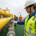 Funcionária Petrobras com colete verde e capacete branco ajusta válvula em tubulação amarela em plataforma marítima.
