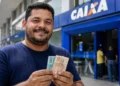 Homem sorridente segurando cédulas de real em frente a uma agência da Caixa, representando o saque de R$ 6.220.