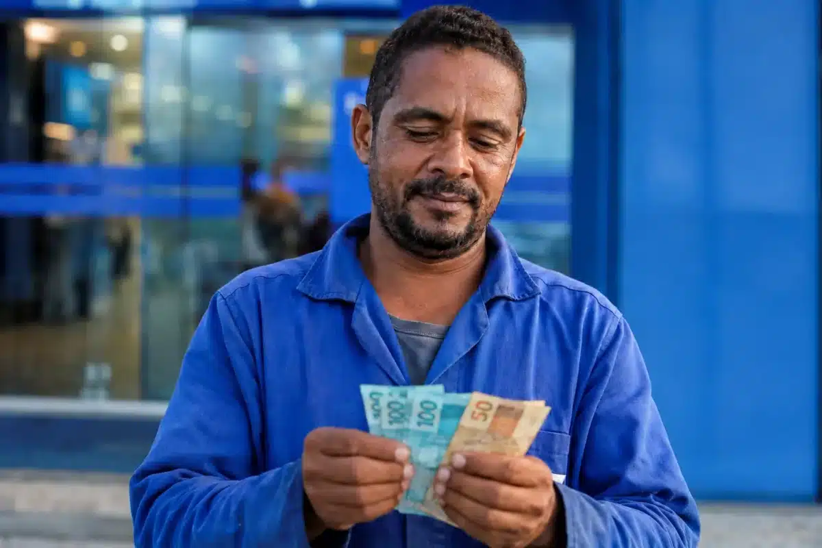 Trabalhador brasileiro conferindo notas de reais em frente a prédio bancário, representando o saque de R$ 6,2 mil do FGTS.