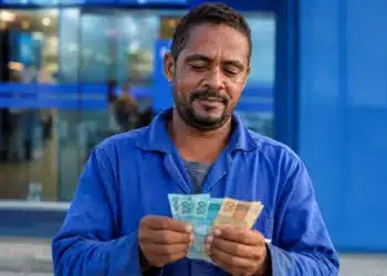 Trabalhador brasileiro conferindo notas de reais em frente a prédio bancário, representando o saque de R$ 6,2 mil do FGTS.