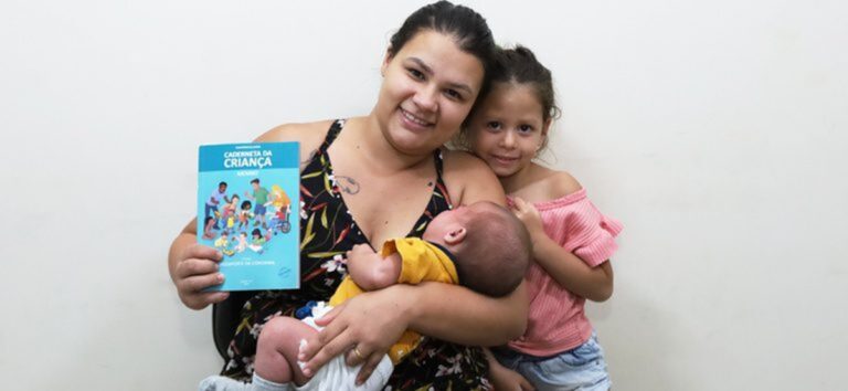 Mãe segura bebê no colo e exibe a Caderneta da Criança ao lado da filha em unidade de saúde