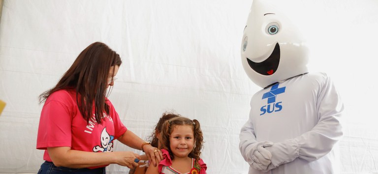 Profissional de saúde vacina menina sorridente ao lado do mascote Zé Gotinha em campanha do SUS