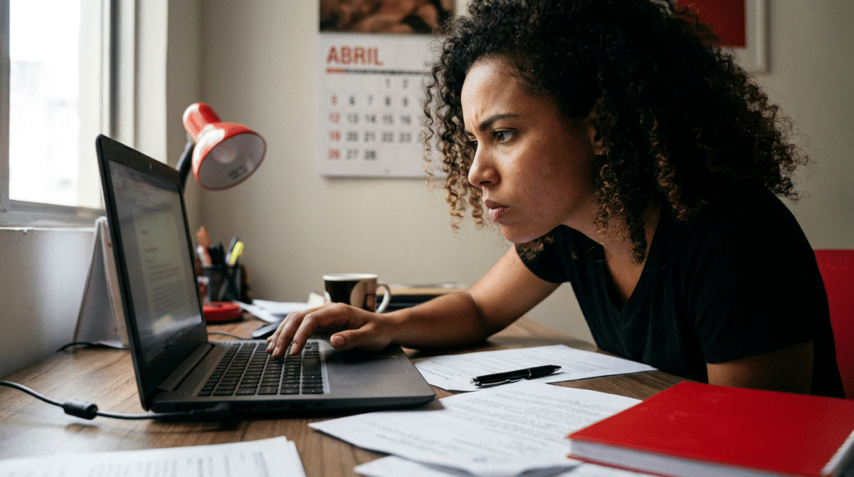 Mulher de cabelos cacheados usando camiseta preta trabalha em laptop sobre mesa com papéis, caneta e luminária vermelha.